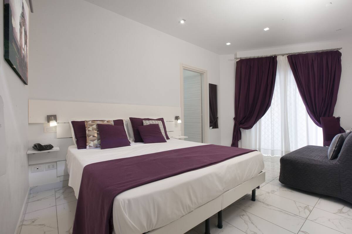 A modern bedroom with a large bed, a bedside table, a nightstand, a sofa, and a window with curtains.