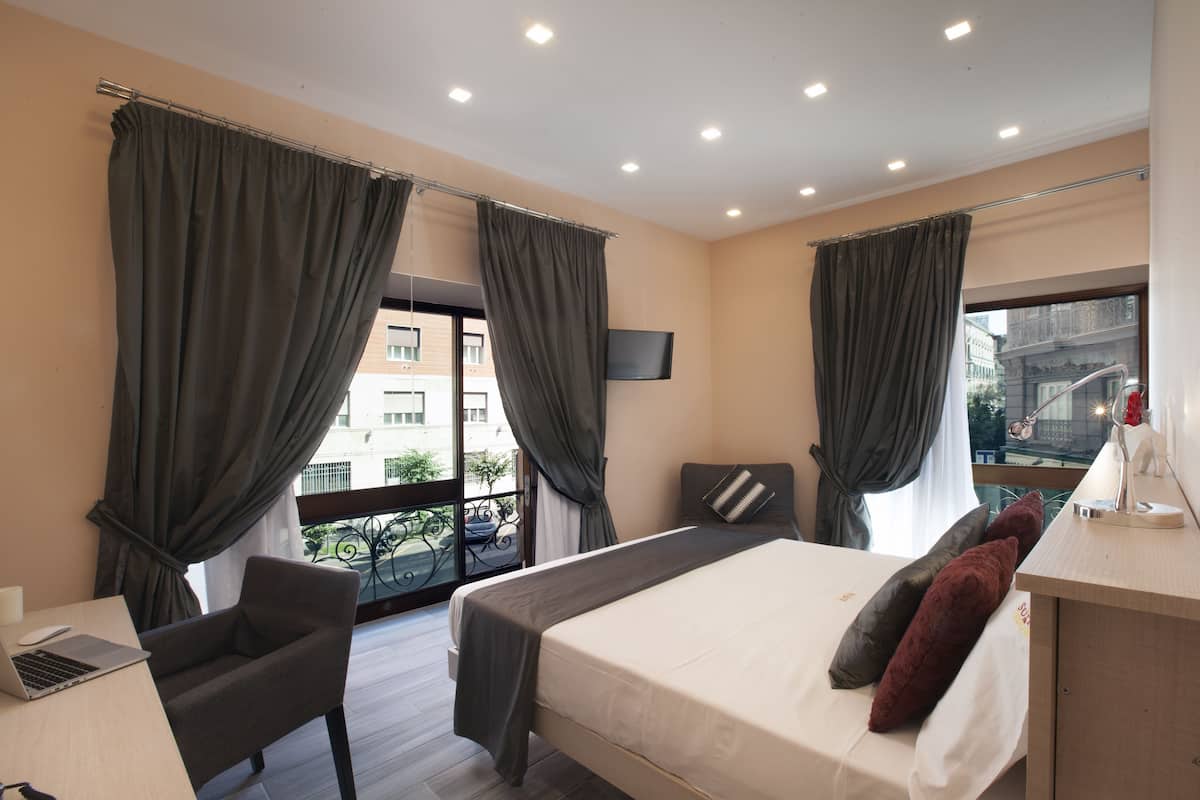 A hotel room with a bed, a desk, a chair, and a balcony with a view of the city.