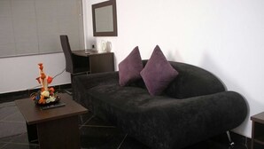 A modern living room with a dark grey sofa, two purple cushions, a small table with a decorative item, a chair, and a desk with a kettle. - PSB Guest House (Accra)