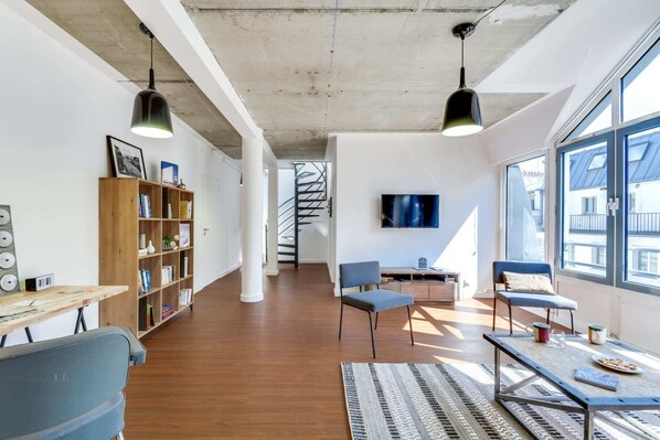 Un espacio habitable amplio con una estantería de madera, un televisor montado en la pared y una escalera que conduce a un piso superior.