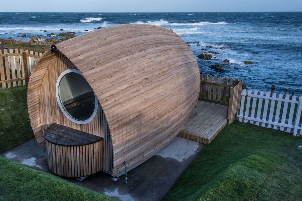 Eine zylindrische Holzkonstruktion mit rundem Fenster und einer kleinen Holzbank, gelegen am Meer mit einem weißen Zaun.