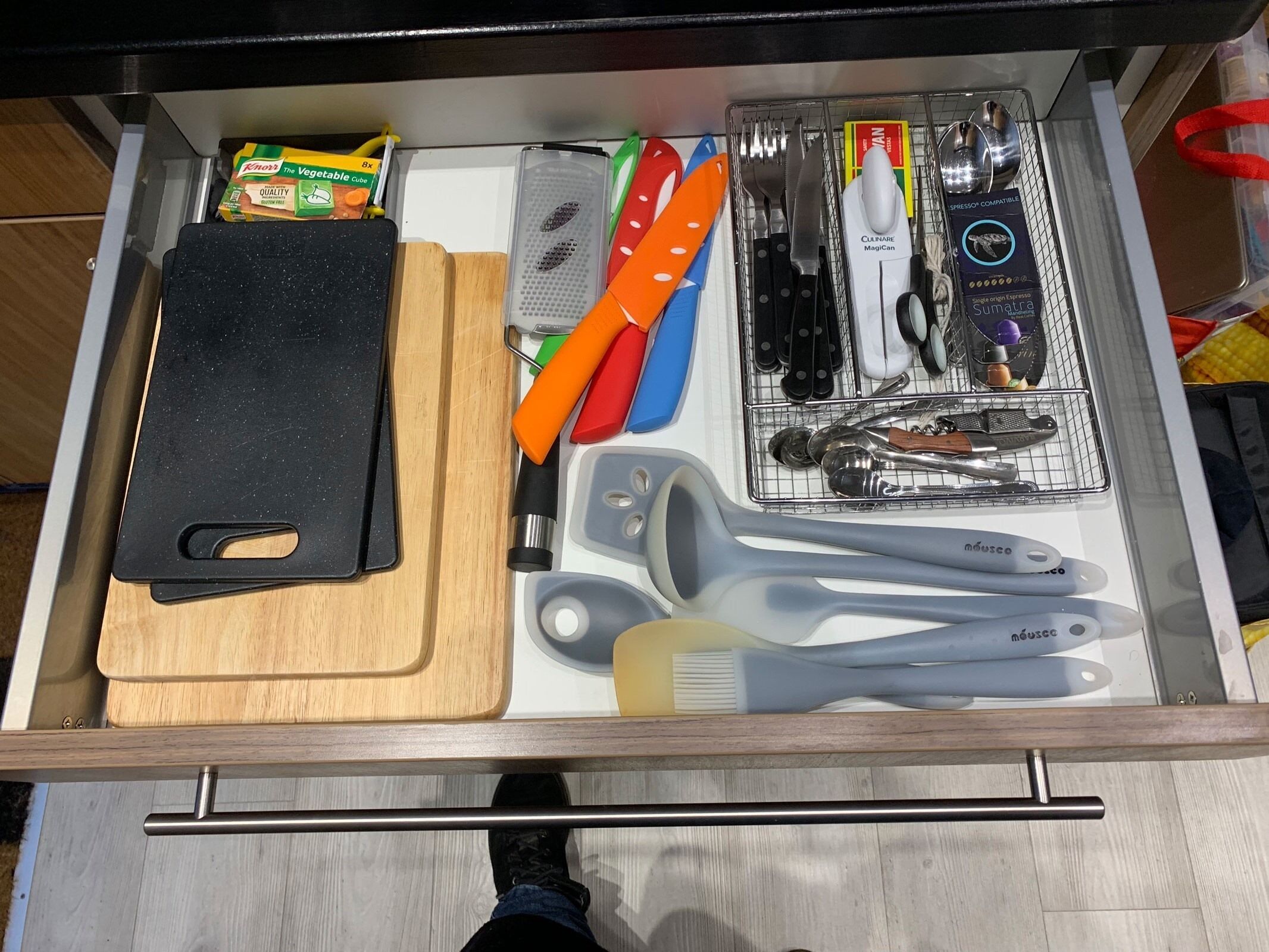 A drawer with a black cutting board, a grater, a set of knives, a set of measuring spoons, and a box of Knorr vegetable cubes.
