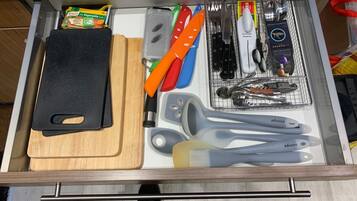 A drawer with a black cutting board, a grater, a set of knives, a set of measuring spoons, and a box of Knorr vegetable cubes.