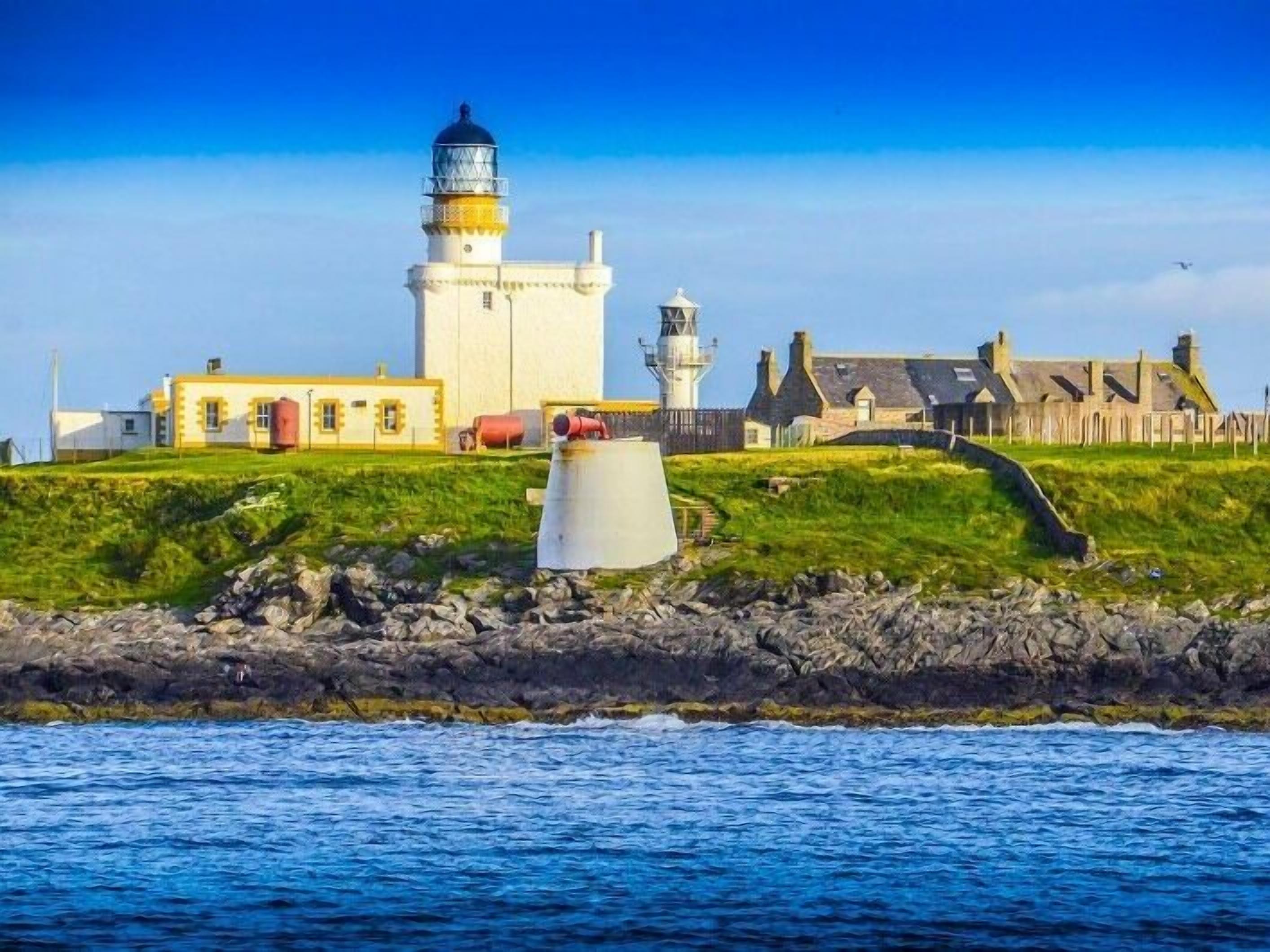 A lighthouse with a white base and a black top, situated on a grassy hill with a rocky shoreline in the foreground.