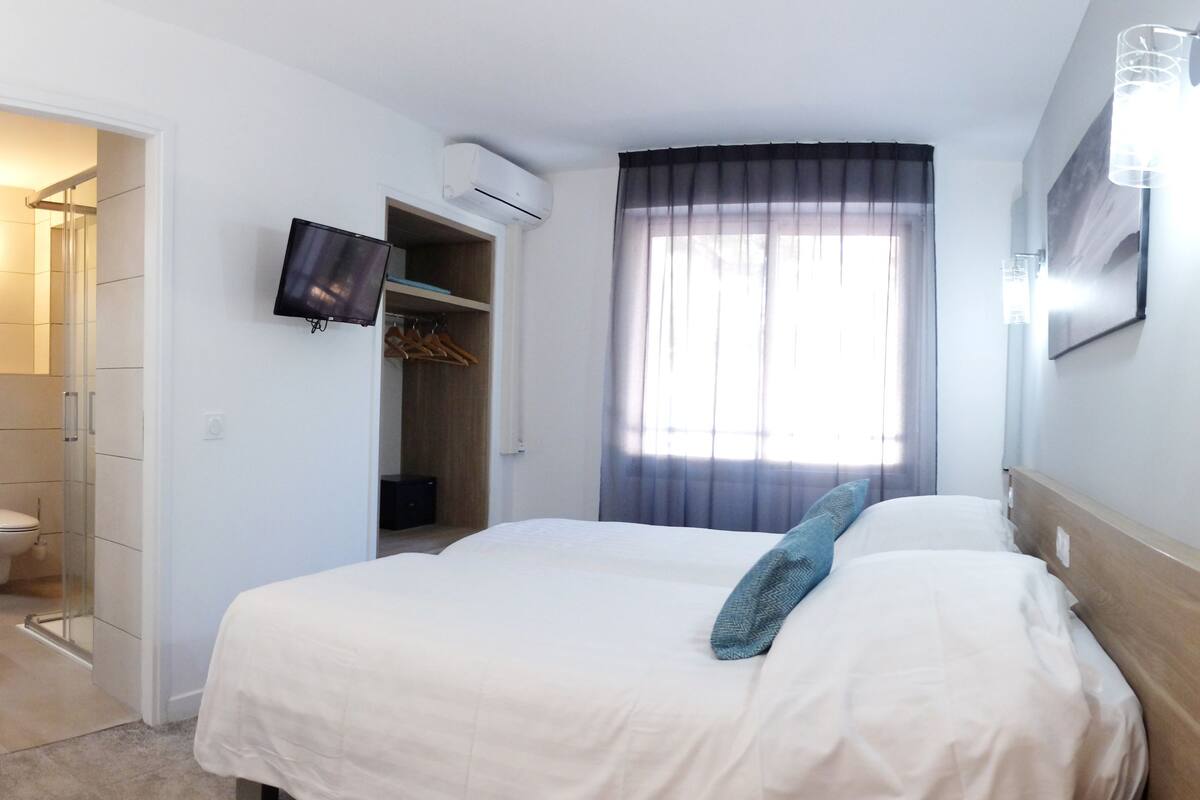 Hotelkamer met een bed, een aan de muur bevestigde televisie, een airconditioningapparaat, een raam met gordijnen en een badkamer met douche en toilet.