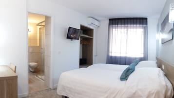 A hotel room with a bed, a TV mounted on the wall, an air conditioning unit, a window with curtains, and a bathroom with a shower and toilet.