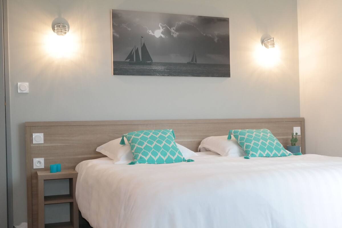 Een slaapkamer met een houten hoofdbord, twee witte kussens, een blauwgroen kussen met patroon, een klein nachtkastje met een blauw object, en een schilderij van zeilboten aan de muur.