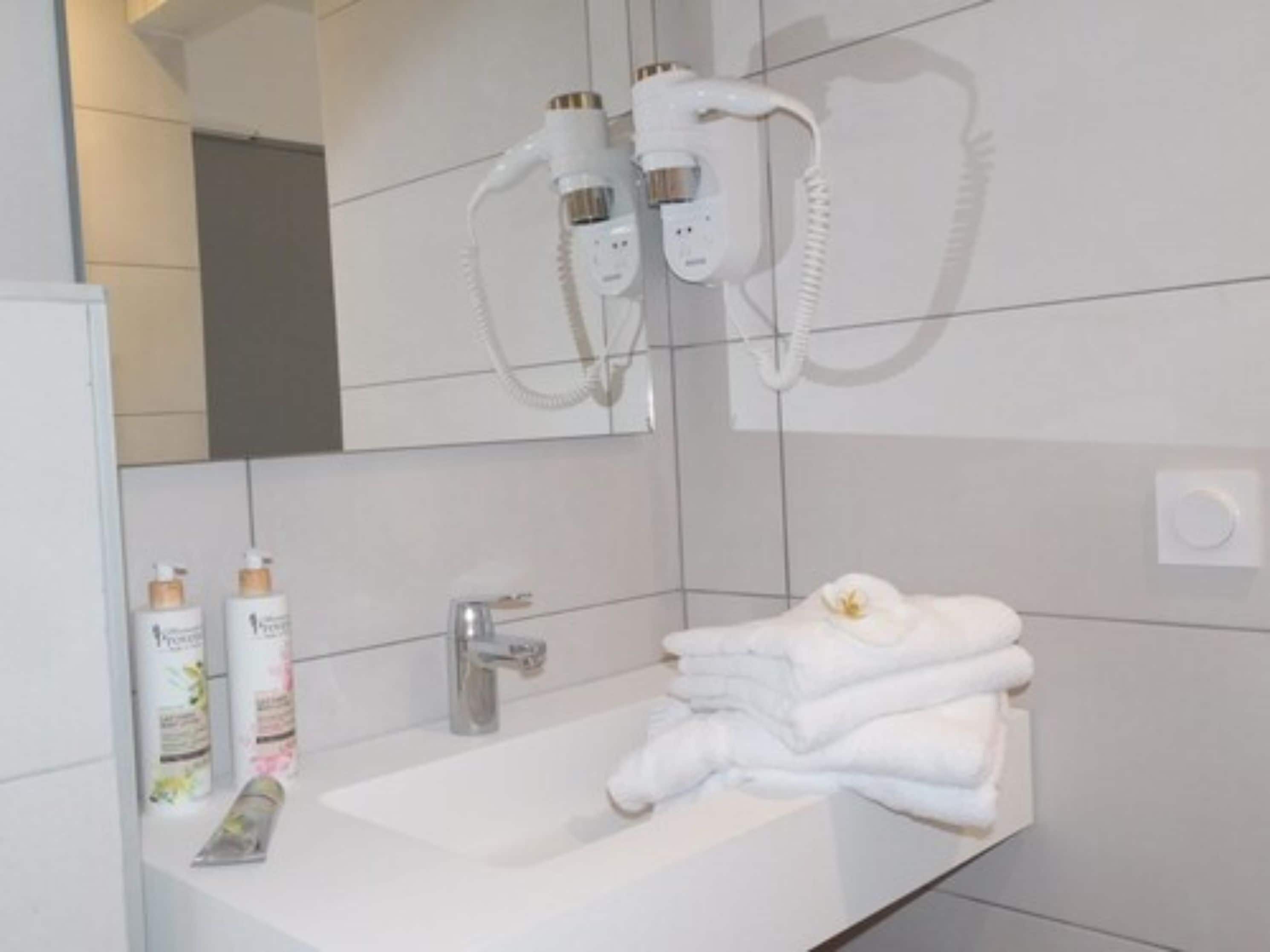 A bathroom with a white sink, two hair dryers mounted on the wall, and a stack of white towels on the counter.