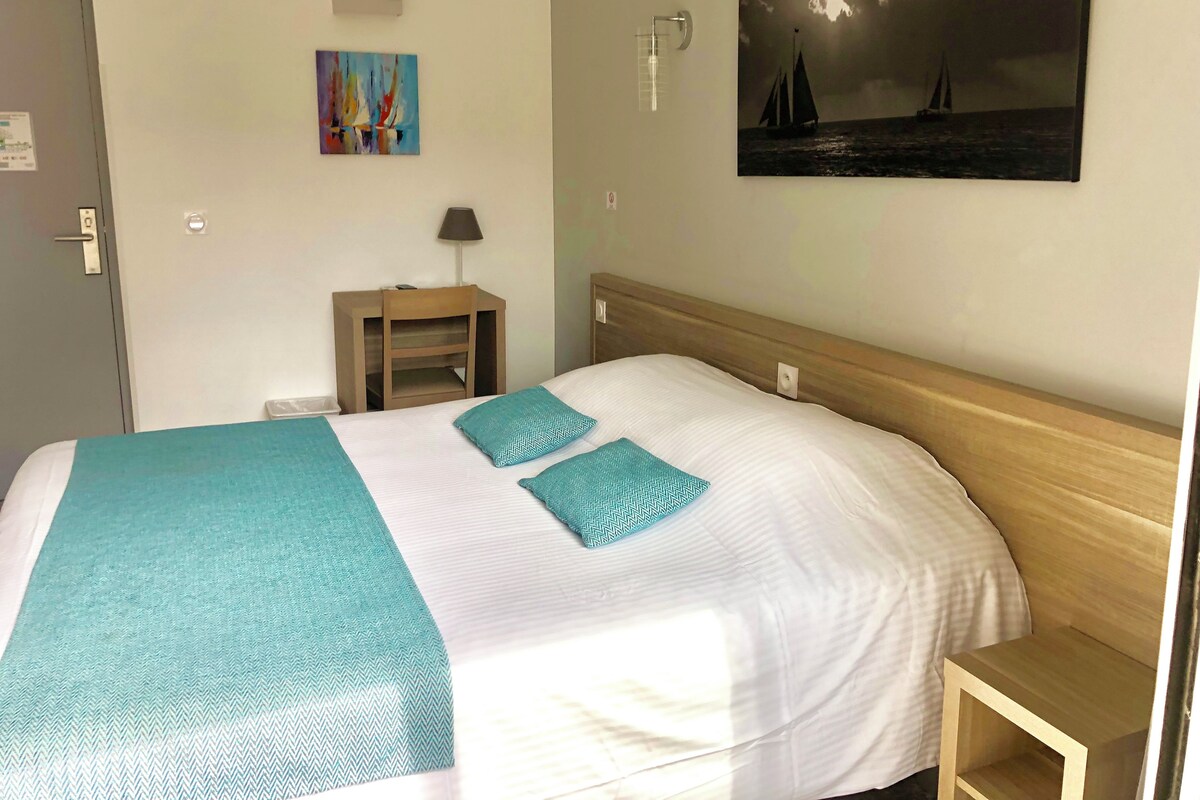 Una cama bien hecha con colcha blanca y azul verdoso, un cabecero de madera, una mesita de noche con una lámpara, y un cuadro de un velero en la pared.