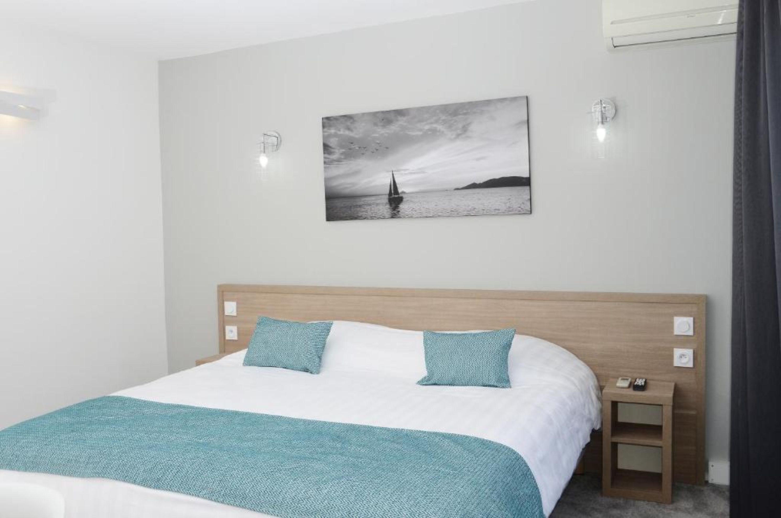 Un dormitorio con una cama, dos almohadas, una mesita de noche y una lámpara de pared, además de un cuadro enmarcado.