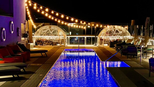 Un espace piscine agrémenté d’un élément décoratif aquatique, de sièges et d’une structure couverte illuminée de guirlandes lumineuses.
