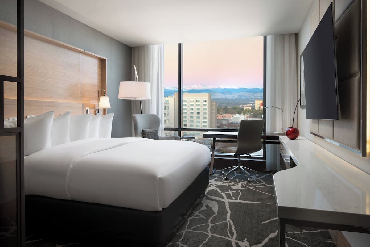 A hotel room with a large bed, a desk, a chair, a TV, and a view of the city and mountains.