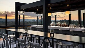 Un bar situé sur un toit, avec un long comptoir, des chaises blanches et une vue sur la ville au coucher du soleil.