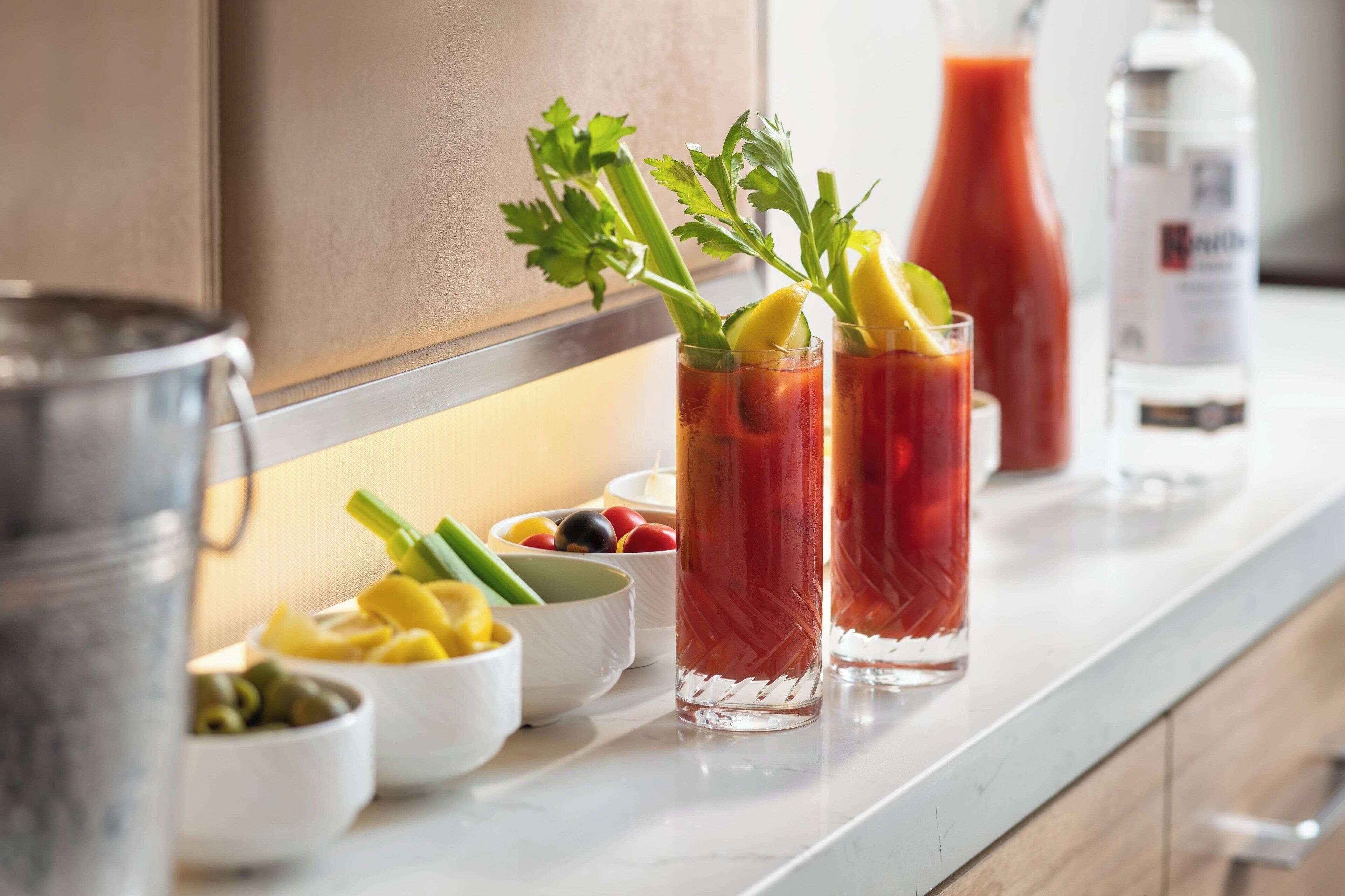 Eine Küchenarbeitsplatte mit zwei Bloody Mary Cocktails, garniert mit Sellerie und Zitrone, einer Tomatensaftflasche sowie Schalen mit Oliven und Essiggurken.