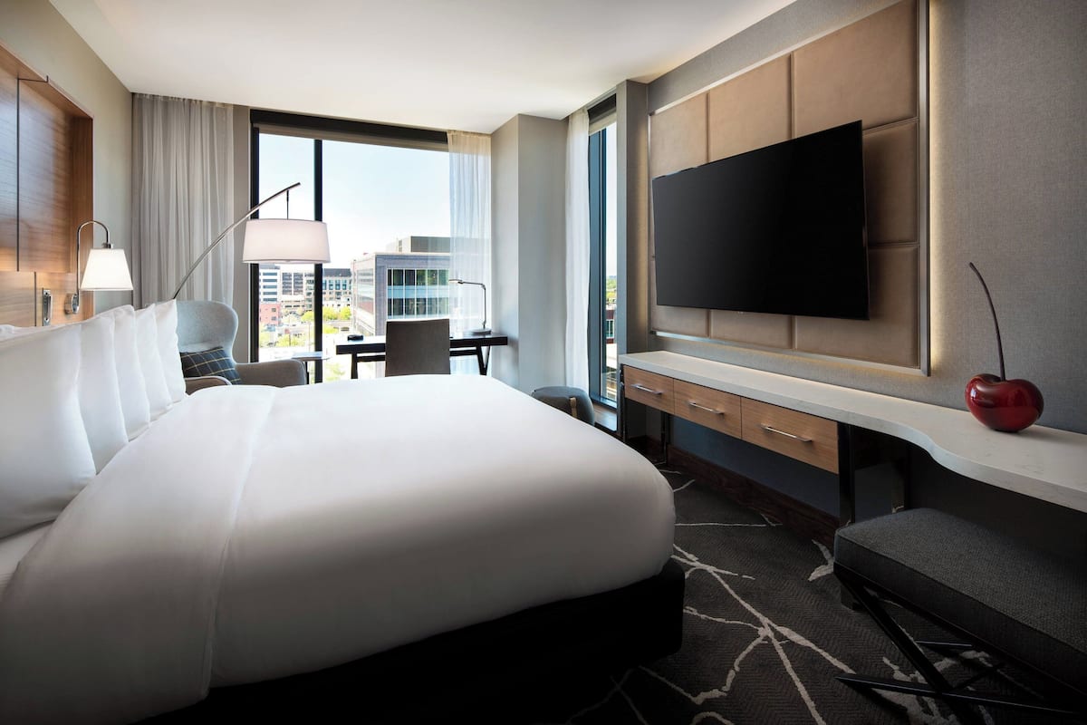 A modern hotel room with a large bed, a desk, a chair, a flat-screen TV, and a view of the city.