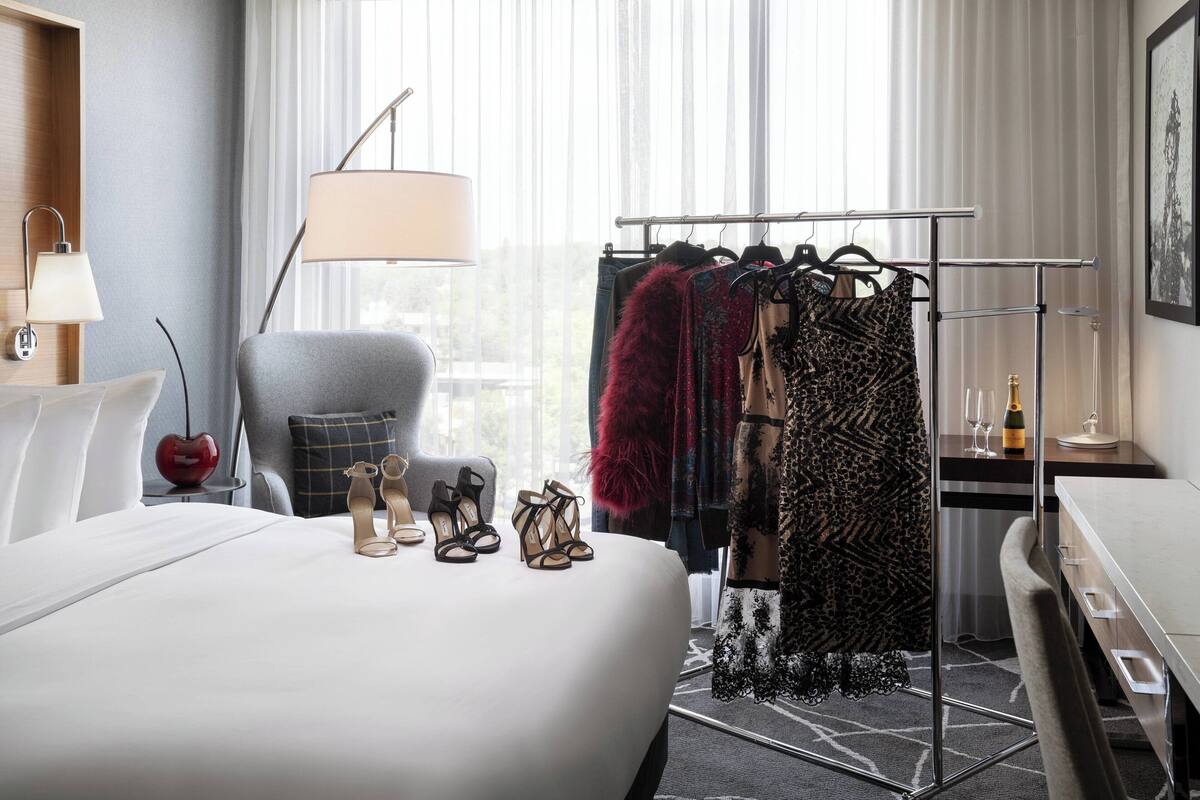 A hotel room with a bed, a chair, a coat rack with clothes, a lamp, and a bottle of champagne.