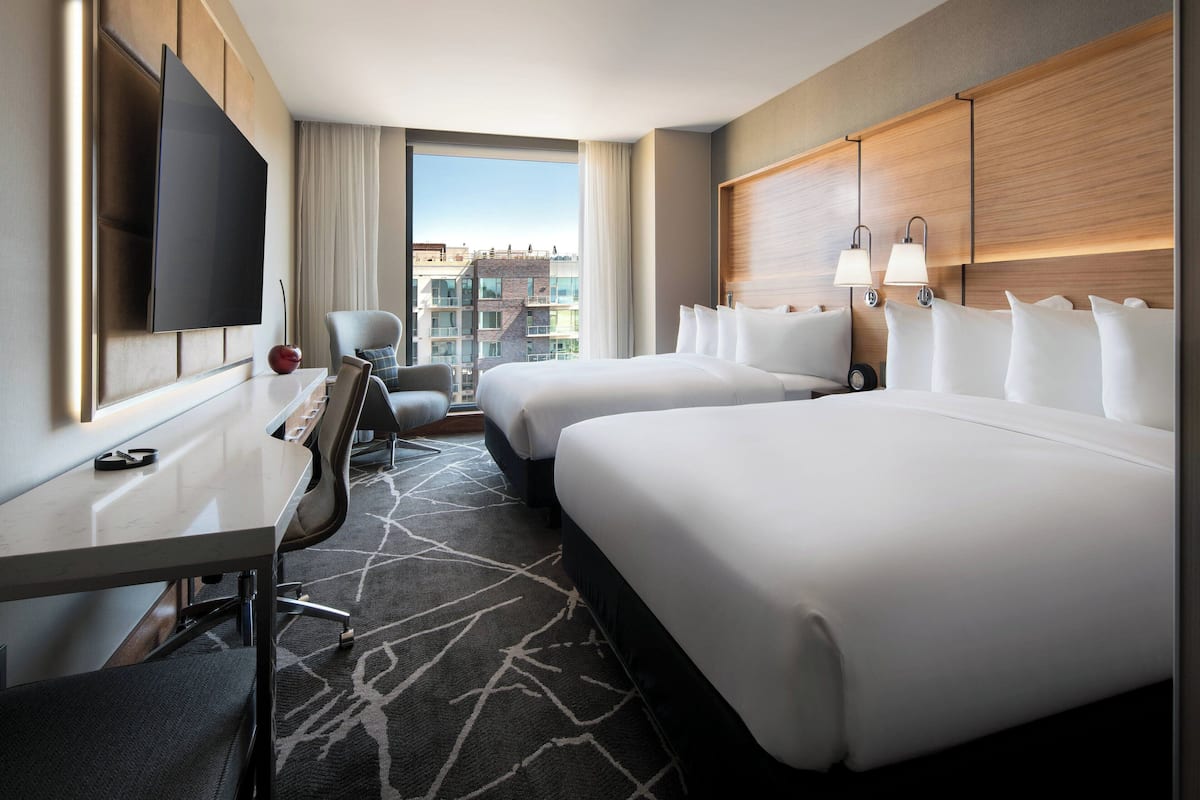 A hotel room with two beds, a desk, a chair, a TV, and a view of the city.