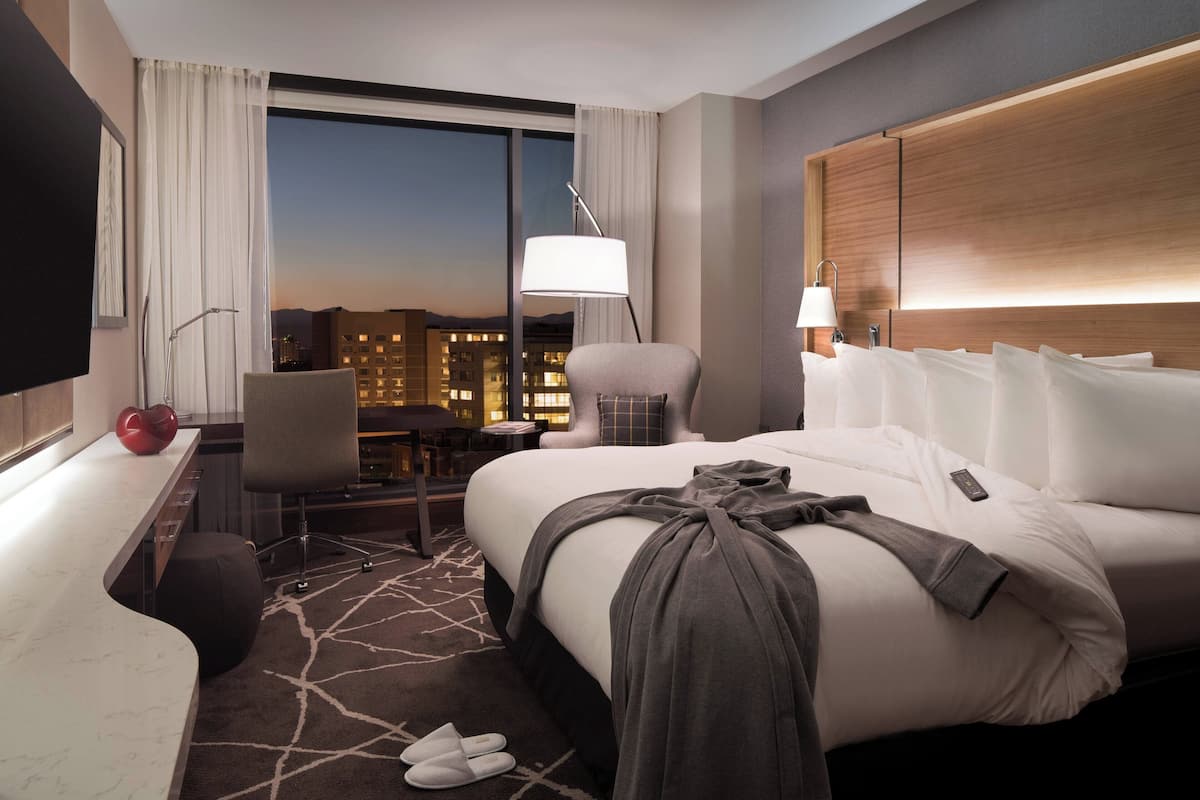 A hotel room with a large bed, a desk, a chair, a TV, and a view of the city at dusk.