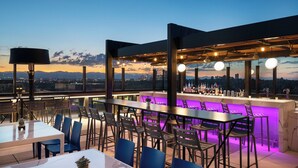 Un espace de restauration en plein air avec des tables et des chaises, un bar avec des bouteilles, et une vue sur la ville au crépuscule.