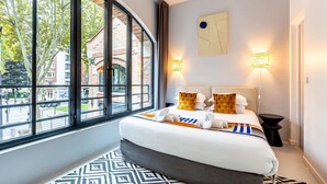 A hotel room with a large window, a bed with white bedding and two pillows, a bedside table, and a patterned rug. - Sweett | Brune VI - Three Bedroom Apartment, Sleeps 8 (Paris)