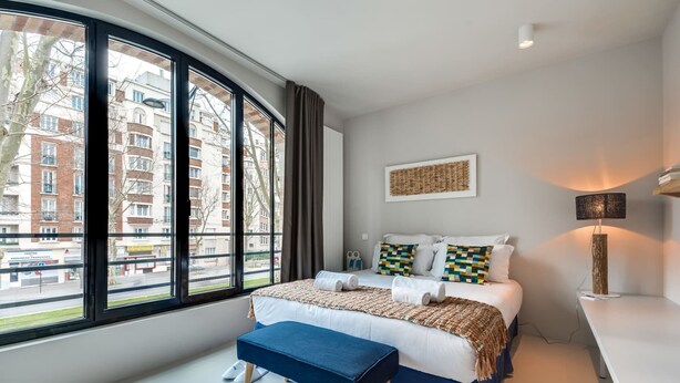 A modern hotel room with a large window, a bed with multiple pillows, a bedside table, and a bench.