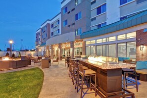 An outdoor bar area with seating and a fire feature, surrounded by a building with multiple floors and large windows. - Courtyard by Marriott Long Island Islip/Courthouse Complex (Central Islip)