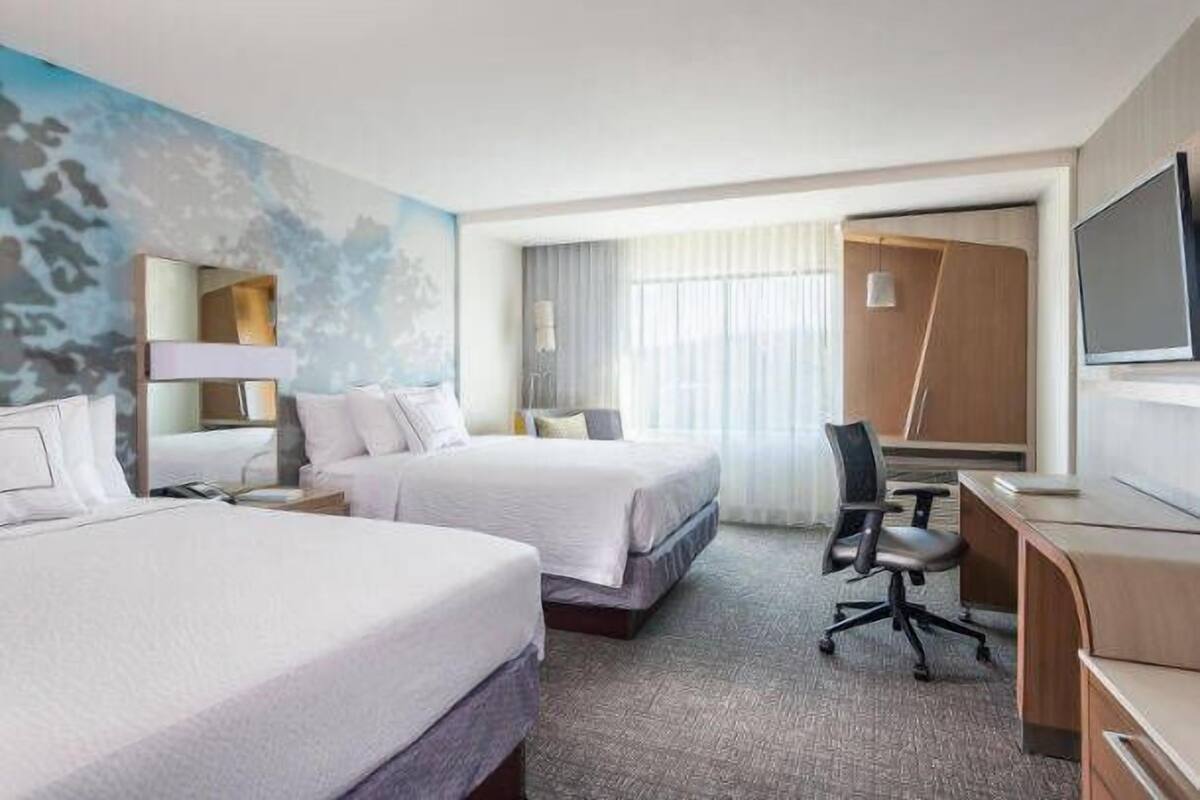 A hotel room with two beds, a desk, a chair, a TV, and a large window with curtains.