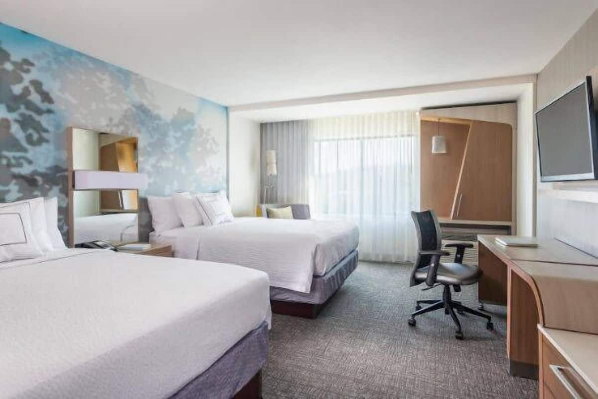 A hotel room with two beds, a desk, a chair, a TV, and a large window with curtains.