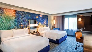 Une chambre d’hôtel avec deux lits, un bureau, une chaise, une grande fenêtre avec des rideaux et une fresque murale colorée.
