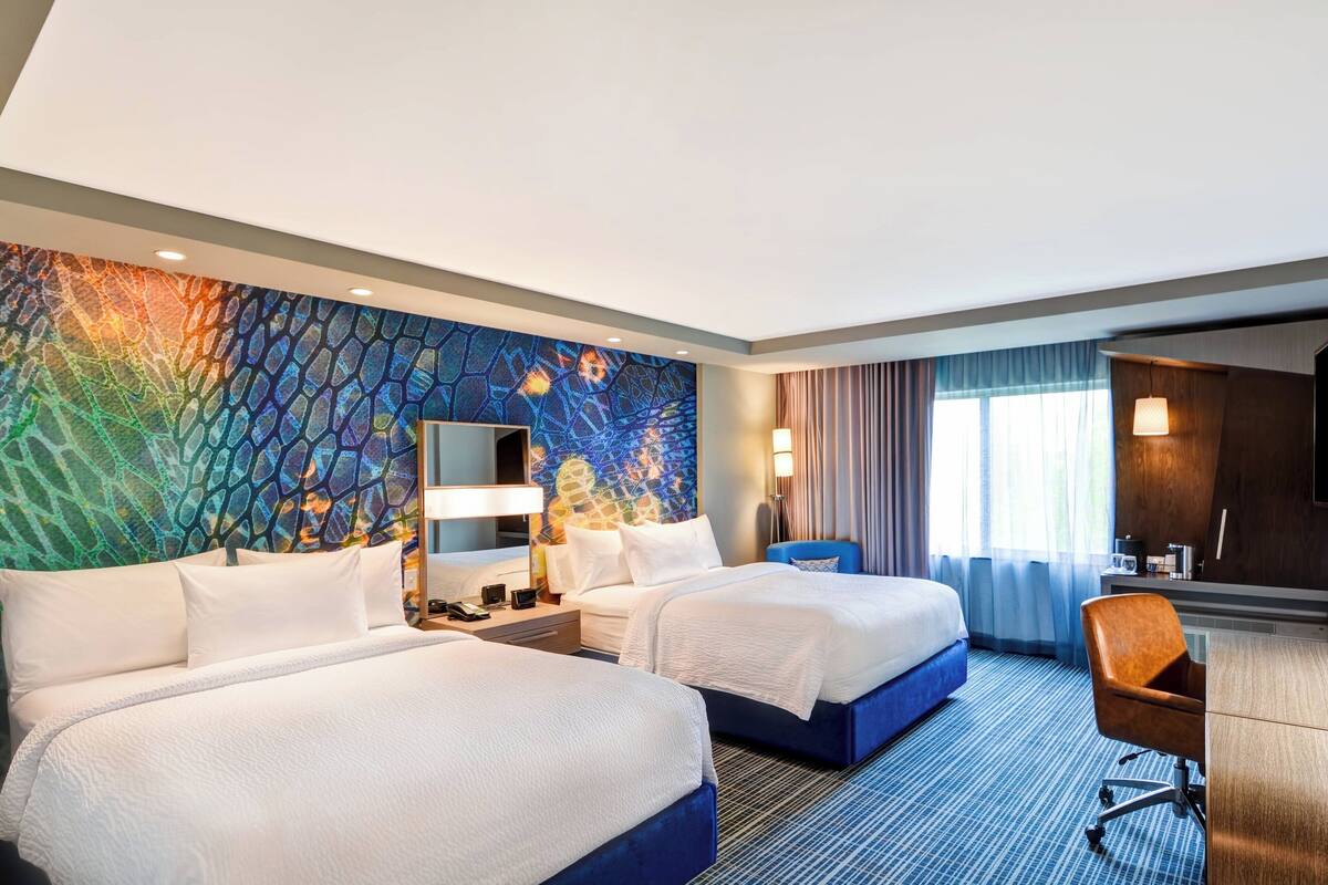 A hotel room with two beds, a desk, a chair, a large window with curtains, and a colorful wall mural.