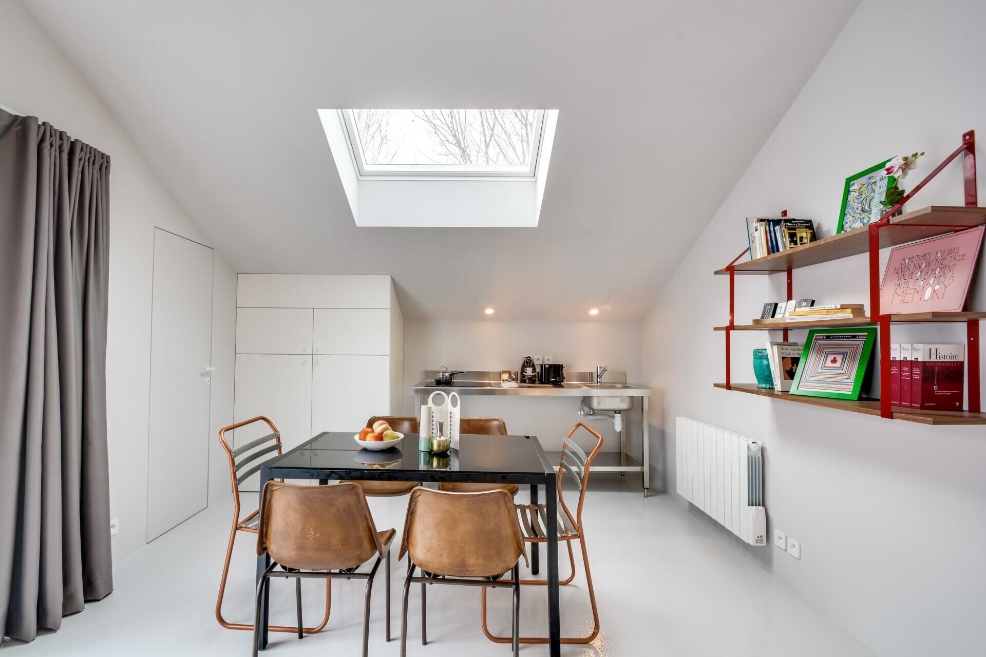 Una cocina moderna con tragaluces, un comedor con mesa y sillas, y una estantería con libros y objetos decorativos.