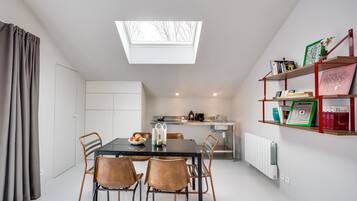 Una cocina moderna con tragaluces, un comedor con mesa y sillas, y una estantería con libros y objetos decorativos.