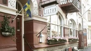 A building with a sign reading "LILTON" and a flag. There are steps leading to the entrance, a bench, and potted plants. - Hotel Lilton (Angelholm)