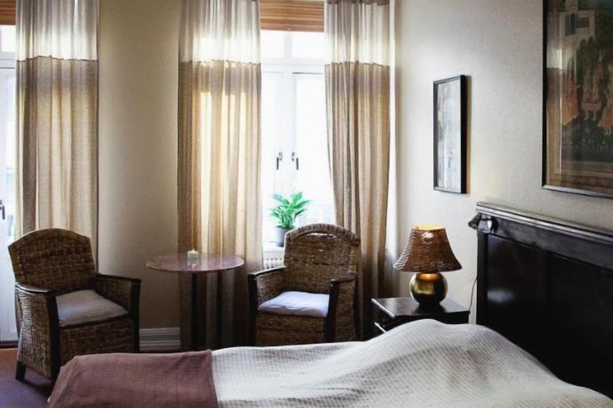 A hotel room with a bed, two chairs, a small table, a lamp, and a painting on the wall.