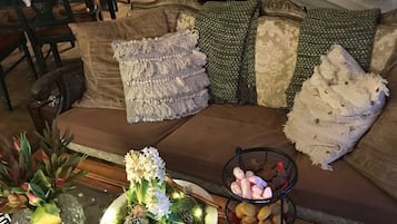 Ett mysigt vardagsrum med en soffa, kuddar, ett soffbord med frukt och en dekorativ skål, samt en matplats med stolar och en kristallkrona.