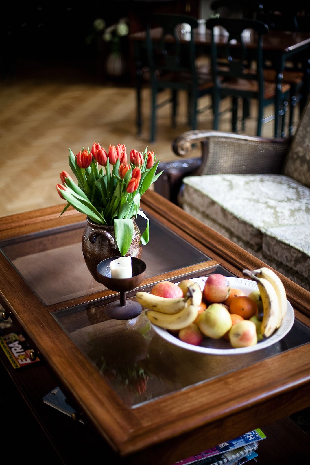 A wooden coffee table with a glass top, a vase of red tulips, and a plate of fruit.