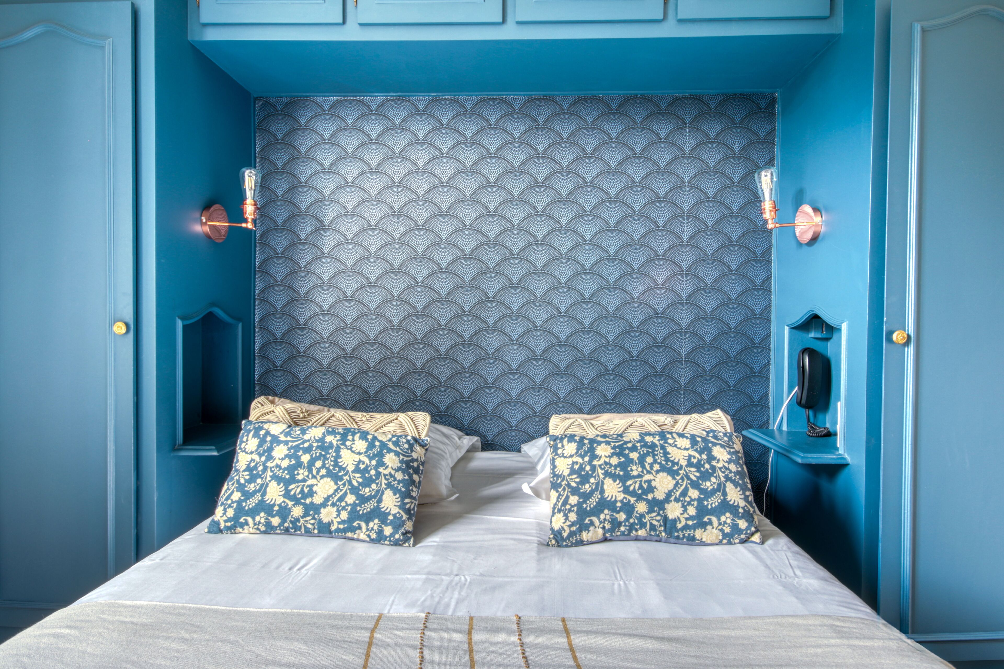 Ein Bett mit zwei Kissen, ein Kopfteil mit geschwungener Musterung und ein Telefon an einer blauen Wand.
