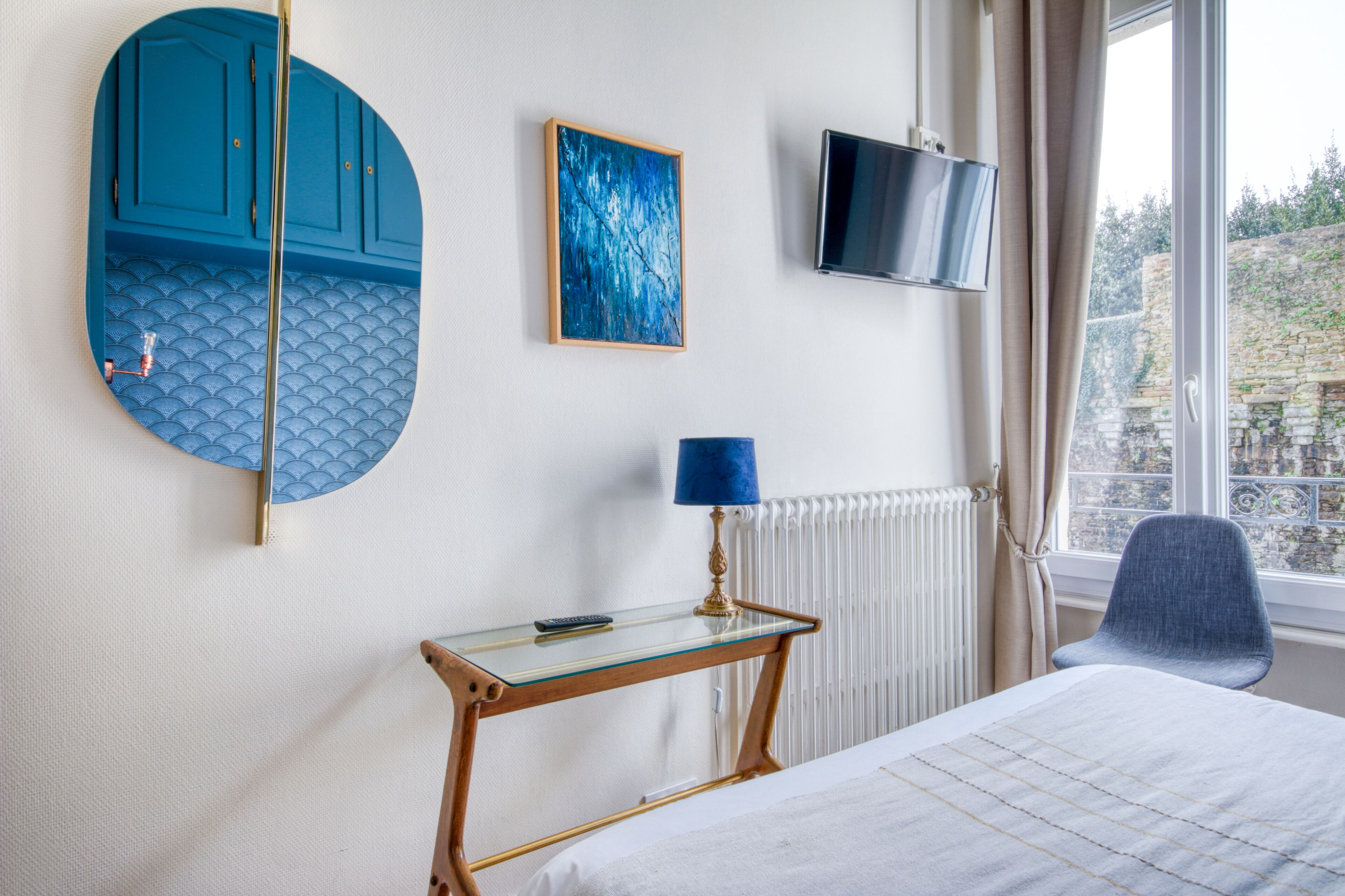 A hotel room with a bed, a round mirror, a TV, a table with a lamp, and a view of a stone wall outside.
