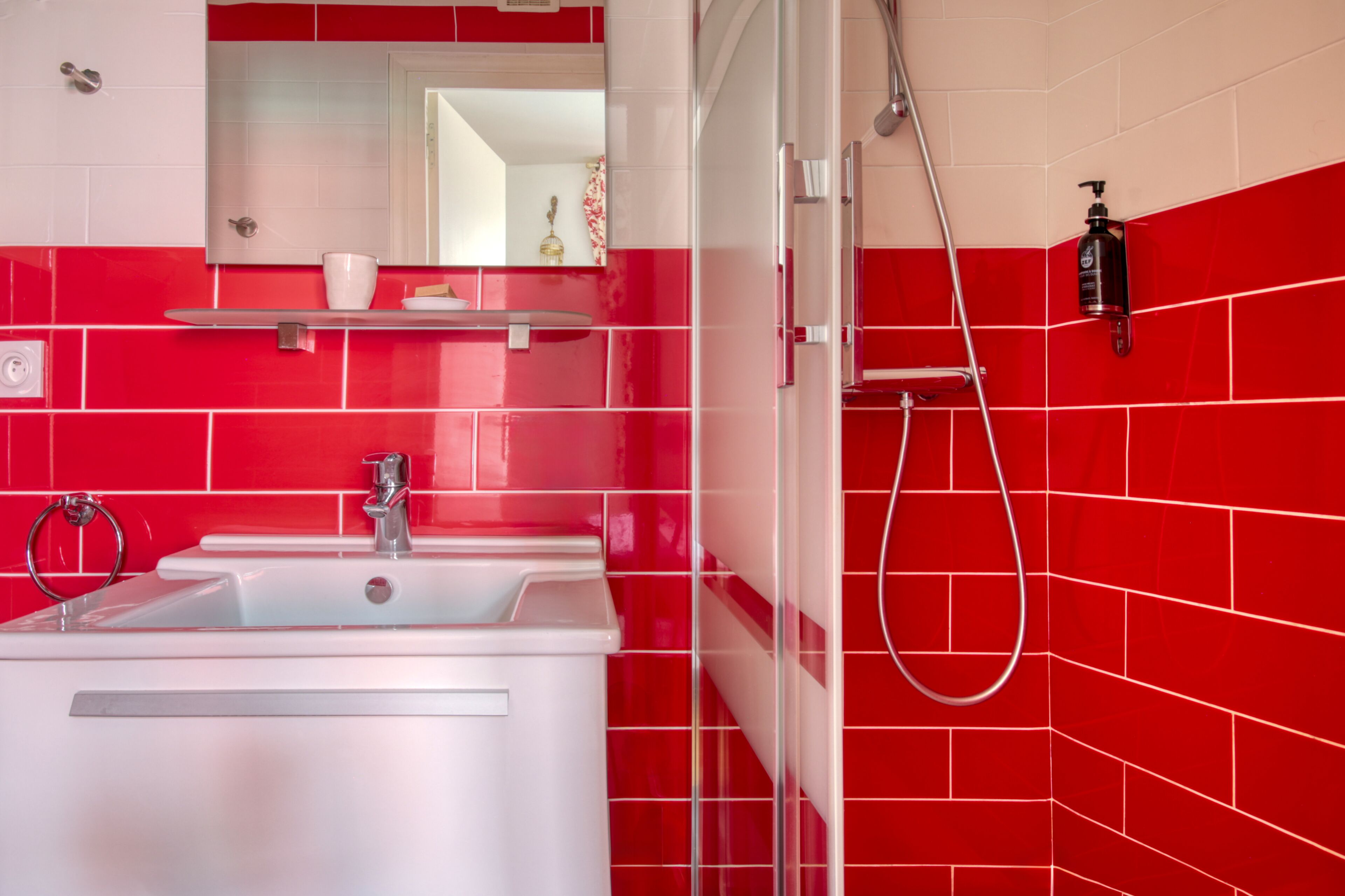 Ein Badezimmer mit einem weißen Waschbecken, einem Spiegel, einer roten Fliesenwand und einer Dusche.