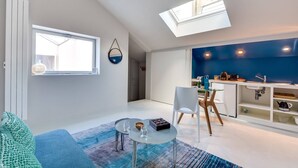 Una sala de estar moderna con una mesa de centro de vidrio, un sofá azul y un comedor con una silla y una mesa blancos. Un tragaluz proporciona luz natural, y se ve una zona de cocina al fondo.