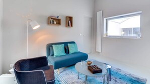 Una sala moderna con un sofá azul, una mesa de centro redonda y una lámpara de pie. Se observan dos estantes con libros y objetos decorativos en la pared.