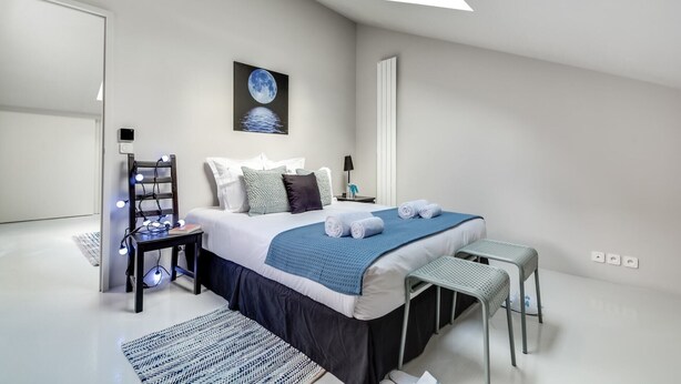 Un dormitorio moderno con tragaluz, una cama con manta azul, una mesita de noche con lámpara y un pequeño taburete.
