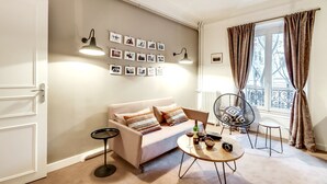 A cozy living room with a sofa, a round coffee table, and a chair. There are wall-mounted picture frames and a window with curtains. - Sweett | Saint Germain I (Paris)