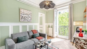 Sala de estar con sofá gris, una mesita de centro, un collage de fotos enmarcadas, un radiador y un balcón con vistas a zonas verdes.