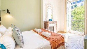 Habitación de hotel con cama, mesita de noche, espejo, chimenea y balcón con vistas a edificios y árboles.