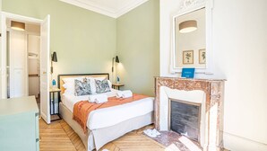 Un dormitorio con cama, lámparas de noche, un espejo, una chimenea y una puerta que da a otra habitación.