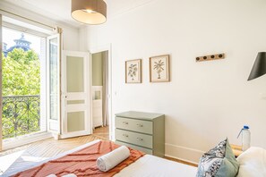 A bedroom with a bed, a dresser, a TV, and a balcony with a view of trees and a building. - Sweett | Saint Germain III (Paris)