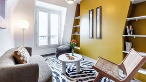 Ein modernes Wohnzimmer mit einer Couch, einem runden Couchtisch, einem Sessel, einem Bücherregal und einem großen Fenster.