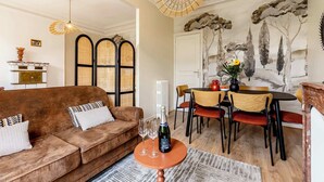 Ein Wohnzimmer mit braunem Sofa, einem Couchtisch und einem Essbereich mit Tisch und Stühlen. Über dem Sofa befinden sich zwei Pendelleuchten und an der Wand hängt ein Baumgemälde.