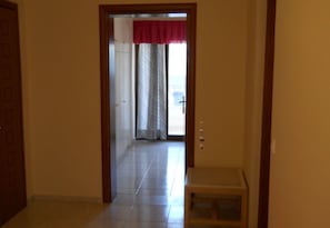 A hallway with a door on the left, a small table on the right, and a view into a room with a window and curtains. - Niriedes Dionyssos Village (Hersonissos)